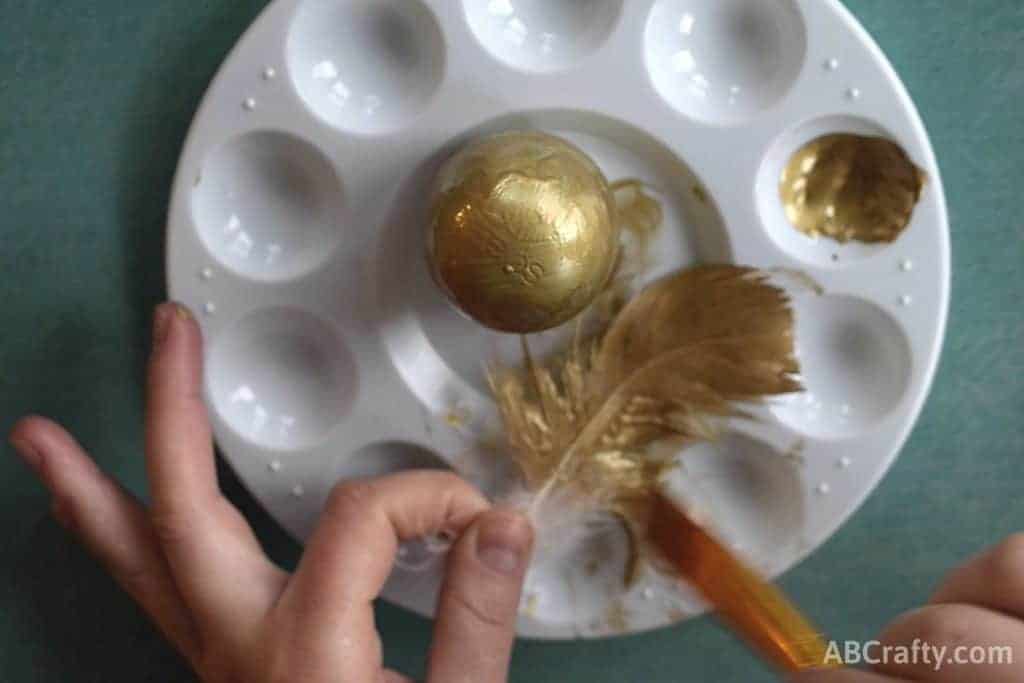 painting a feather with gold paint with an already painted lip balm as part of a harry potter craft to make a golden snitch