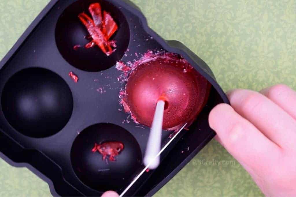 Using a butter knife to break off the extra candy from around the red homemade tootsie pop while still in the silicone mold