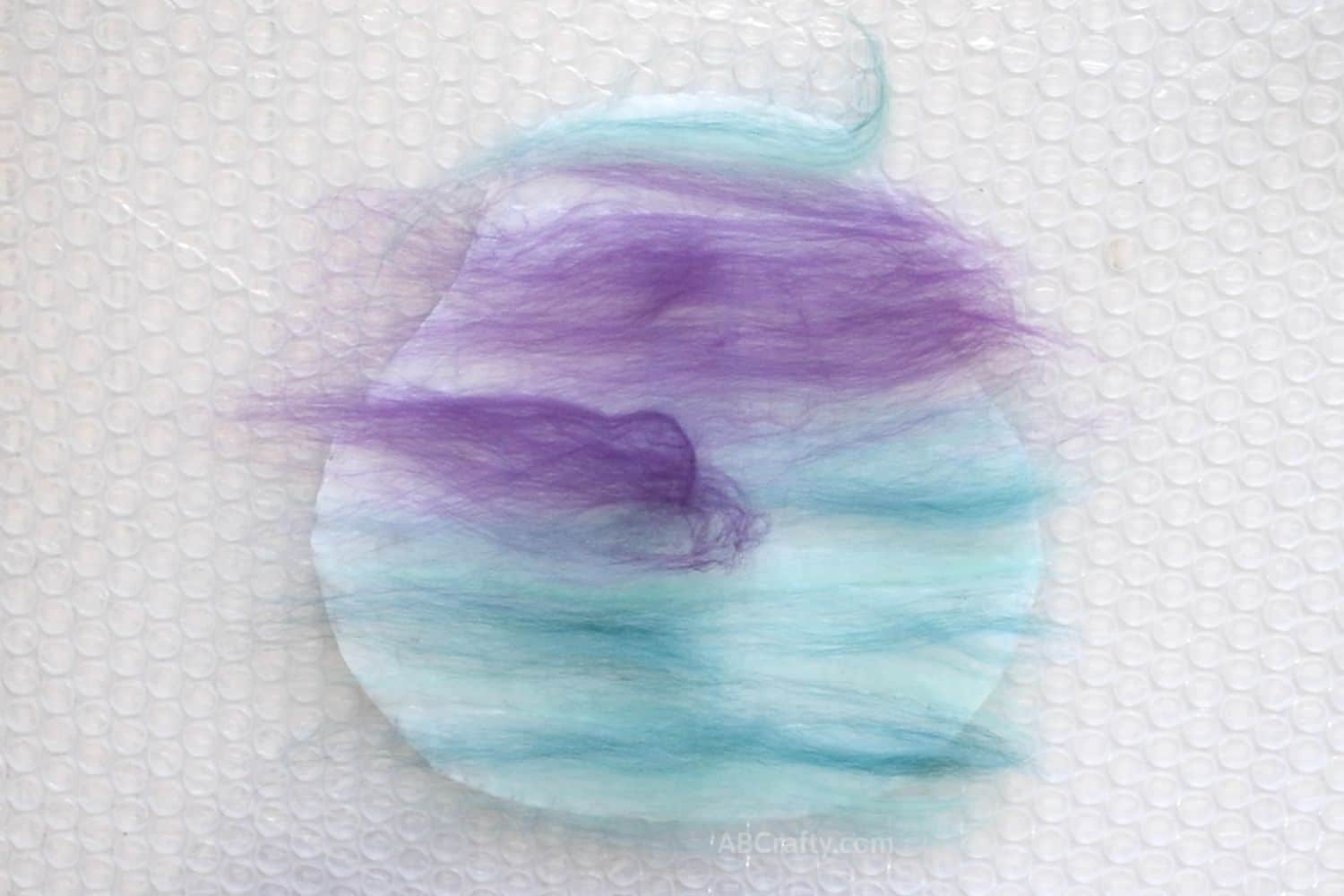 Layer of purple and teal merino wool facing one direction on top of a resist with some wool hanging off the edge