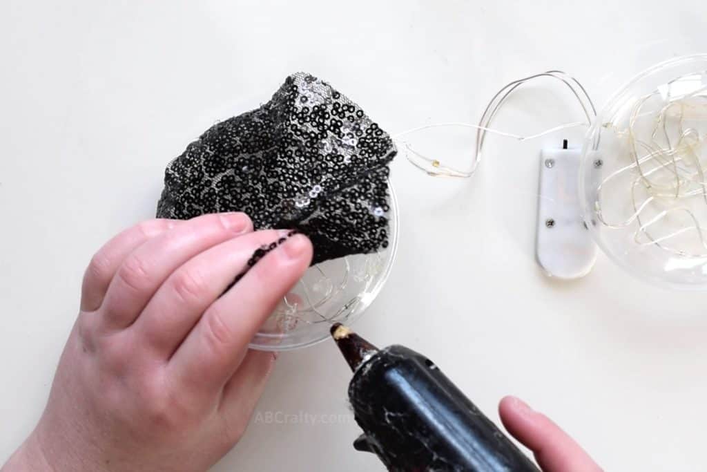 Using a glue gun to glue a circle of black sequin fabric onto a plastic ornament filled with fairy lights