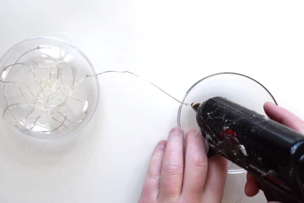 Using a glue gun to glue a wire with LED lights onto half of a fillable plastic ornament while the rest of the fairy lights are already glued into a closed ornament