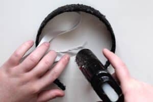 Using a glue gun to glue the sides of a white satin ribbon down. The ribbon is also attached to a black sequin headband.