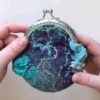 Holding a wet felted coin purse that is purples and greens and has rainbow silk and blue and teal silk ruffles with a metal purse clasp
