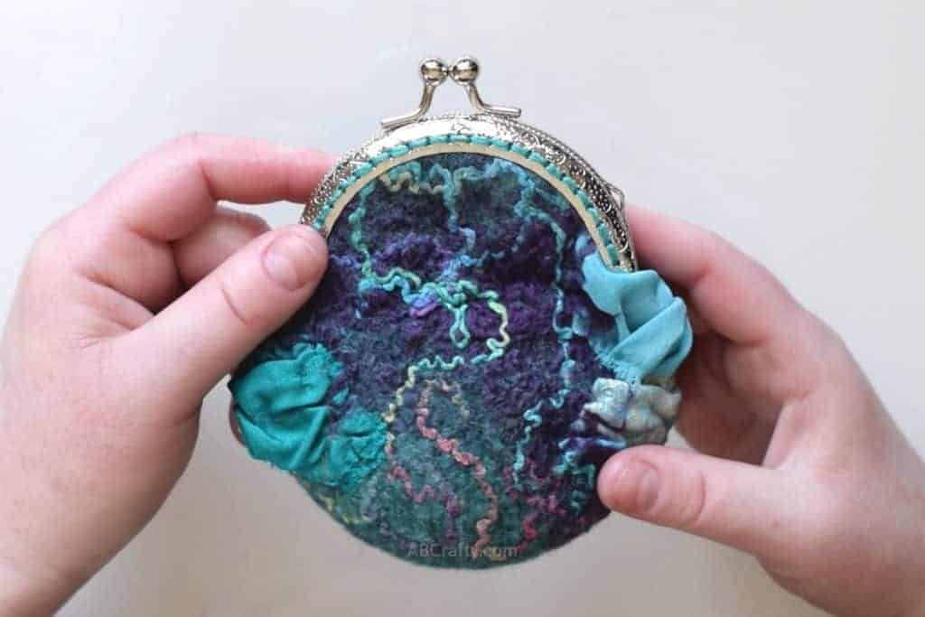 Holding a wet felted coin purse that is purples and greens and has rainbow silk and blue and teal silk ruffles with a metal purse clasp