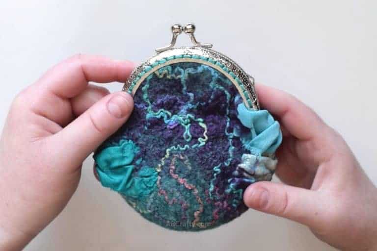 Holding a wet felted coin purse that is purples and greens and has rainbow silk and blue and teal silk ruffles with a metal purse clasp