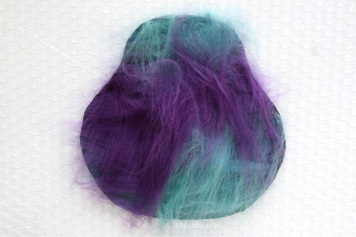 Purple and teal layers of wool fiber on a resist in a coin purse pattern