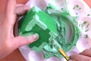 Using paintbrush to paint all corners of painted green cube with stripes of light green fabric paint