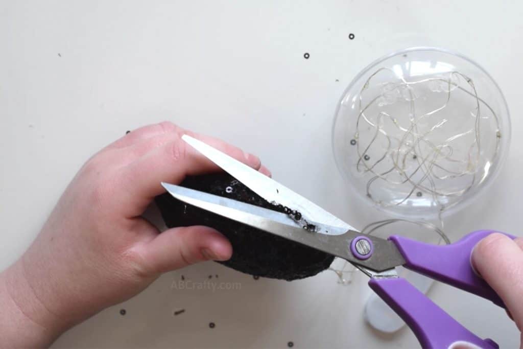 Trimming black sequin fabric glued to an plastic ornament using purple scissors