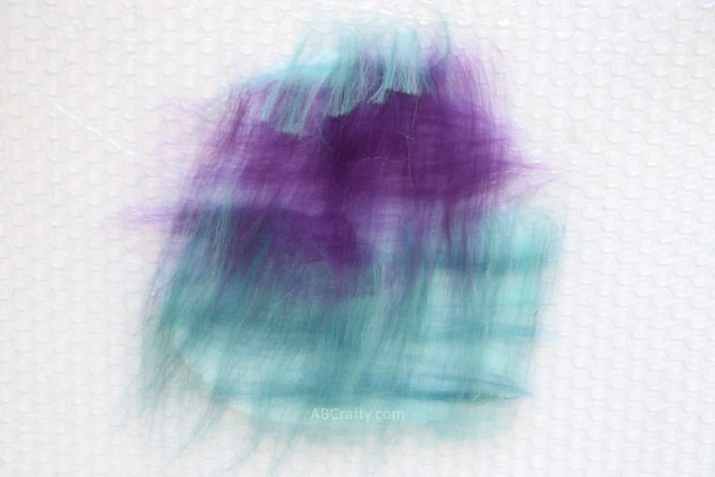 Two perpendicular layers of purple and teal wool on a resist with wisps hanging off the sides