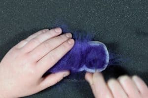 Using a felting needle to poke into purple wool inside of a cookie cutter in the shape of a marshmallow peep