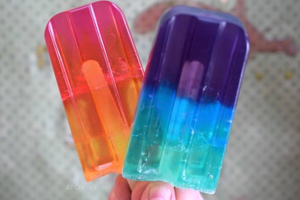 Holding colorful soaps that look like popsicles. One is pink orange and yellow, and the other is purple blue and green