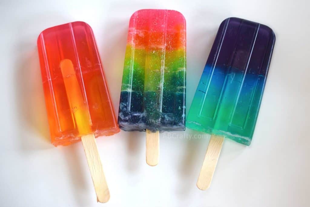 DIY Rainbow Popsicle Soap showing three soaps, 1 in warm colors of pink orange and yellow, rainbow glitter soap, and cool colors with purple blue and green soap. They all have popsicle sticks