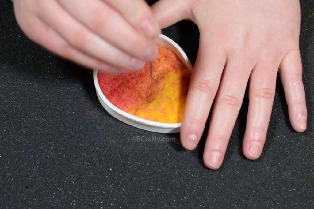 Using a felting needle to felt colorful wool that is pink, orange, and yellow inside of an egg shaped cookie cutter