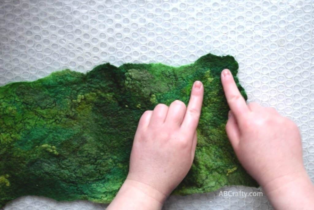 Pointing at the corner of partially wet felted green wool fabric with fun texture and designs from other green fibers