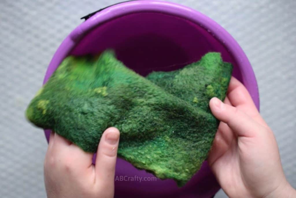 Holding green wet felted wool fabric over a plastic purple bucket filled with water