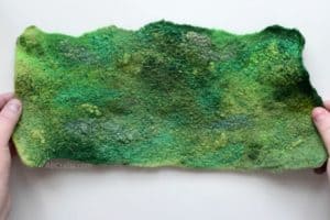 Holding finished piece of handmade wet felted green fabric with swirls of viscose, wool nepps and other embellishment fibers that have been felted into the wool