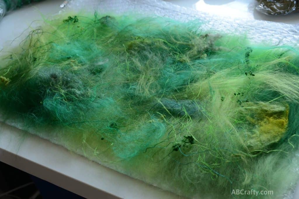 Various green fibers including viscose, ramie, wool nepps, viscose thread, and throwster silk laid out on top of green merino wool on top of bubble wrap