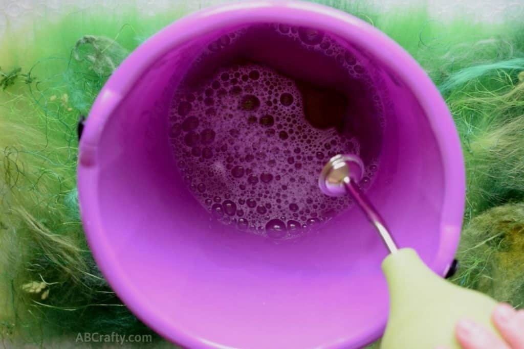 Stirring hot water and olive oil soap in a purple bucket with a green ball brause