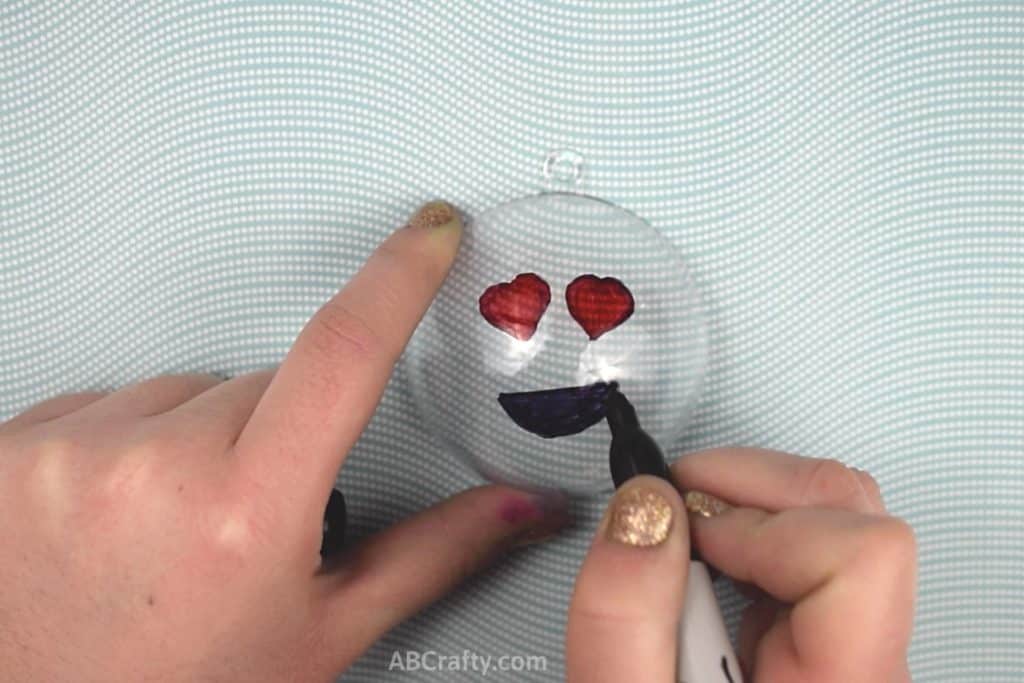 Drawing a black smile onto a heart eyed emoji with a sharpie on top of a plastic ornament