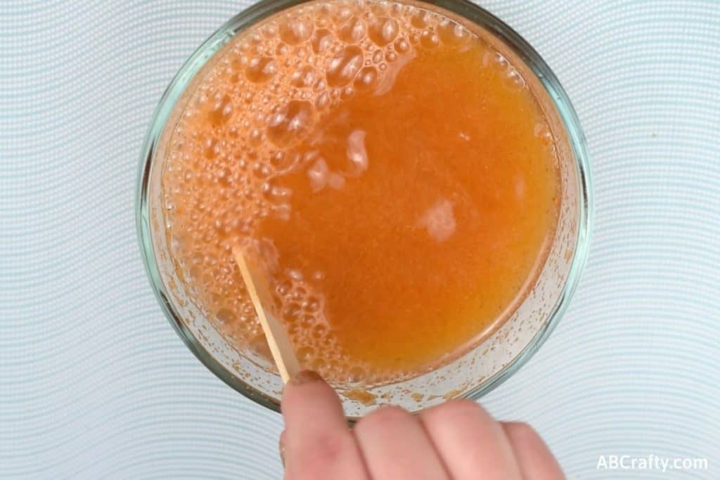 Stirring an orange mixture in a glass bowl with a popsicle stick