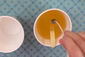 Dipping the metal part of the bottom of a waxed wick into melted wax inside of a paper cup with a popsicle stick in it