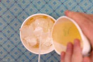 Pouring melted wax out of a paper cup on top of ice cubes with a candle wick sticking out of the middle of a paper cup.