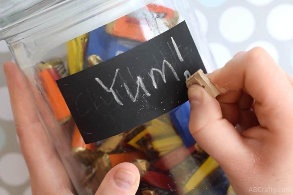 Using a piece of chocolate chalk to write 'yum' on a chalkboard label on a glass jar filled with hershey's miniature chocolate bars