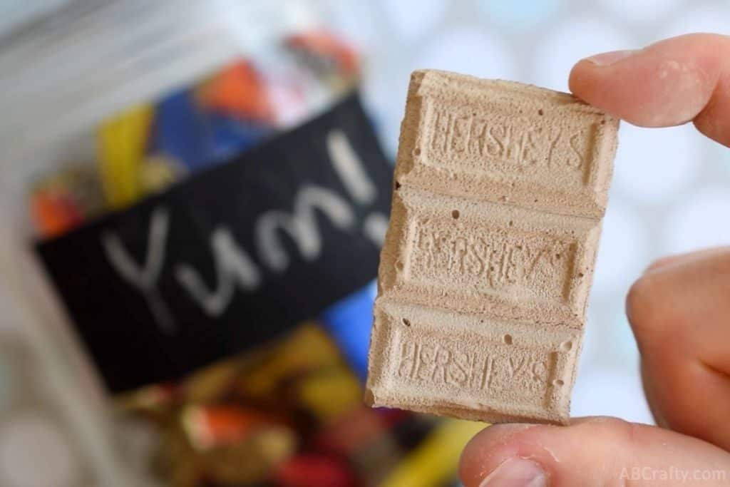 holding a small piece of diy chocolate chalk with a glass jar in the background that has a chalkboard label with 'yum' written on it