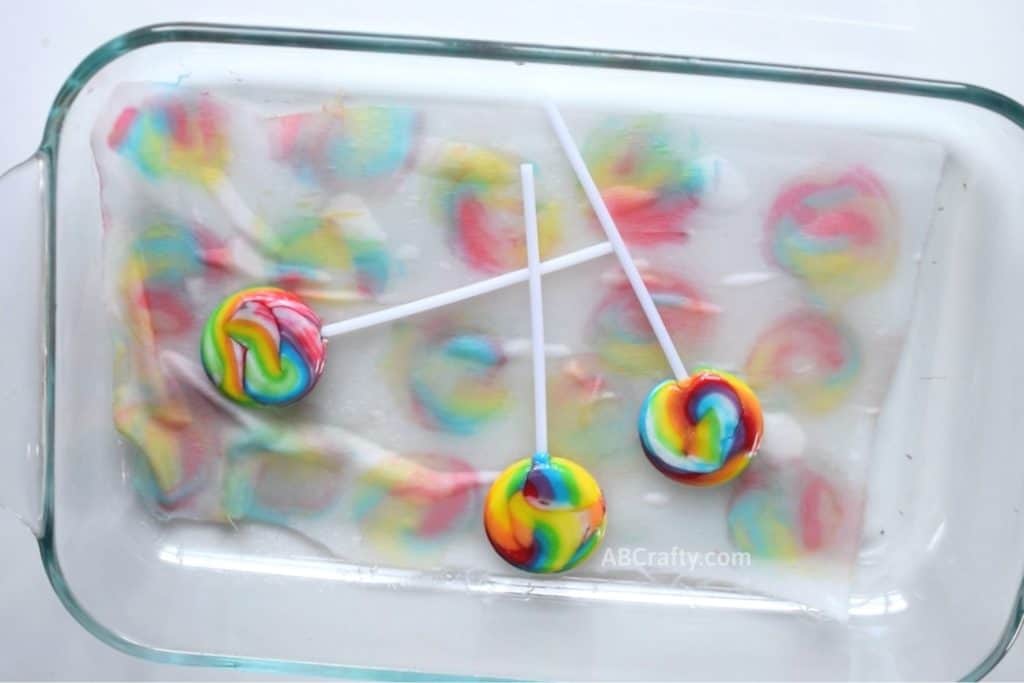 Three lollipops with the food dye partially removed on top of satin fabric, dyeing the satin fabric underneath with swirly colorful circles