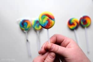 Holding a small round rainbow swirl lollipop with other unwrapped and not unwrapped swirly pops in the background on a white table