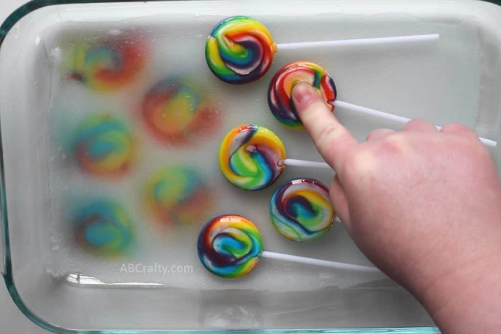 Pressing on a swirly rainbow sucker that has some of the dye gone revealing the white underneath. It's being pressed into a white fabric scrap inside of a pyrex dish with rainbow tie dye swirls made from the lollipops