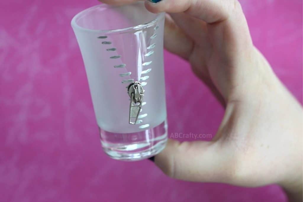 Holding a handmade custom shot glass with an unzipped zipper design painted with silver glass paint and frosted glass with a zipper pull attached