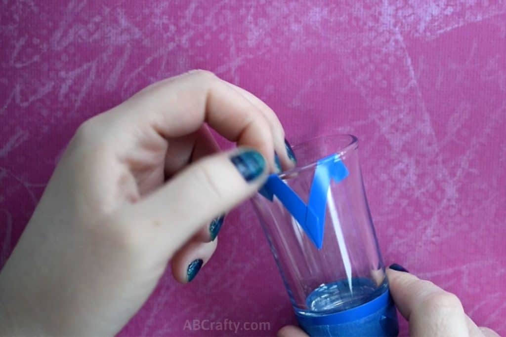 Placing blue vinyl tape at the top of a shot glass to form a stencil in the shape of a triangle