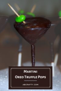 Martini glass cake pop made of oreo balls. It's on a cake pop stick, covered in chocolate with a green jelly bean as the olive on a toothpick. Title reads 'martini oreo truffle pops, abcrafty.com'