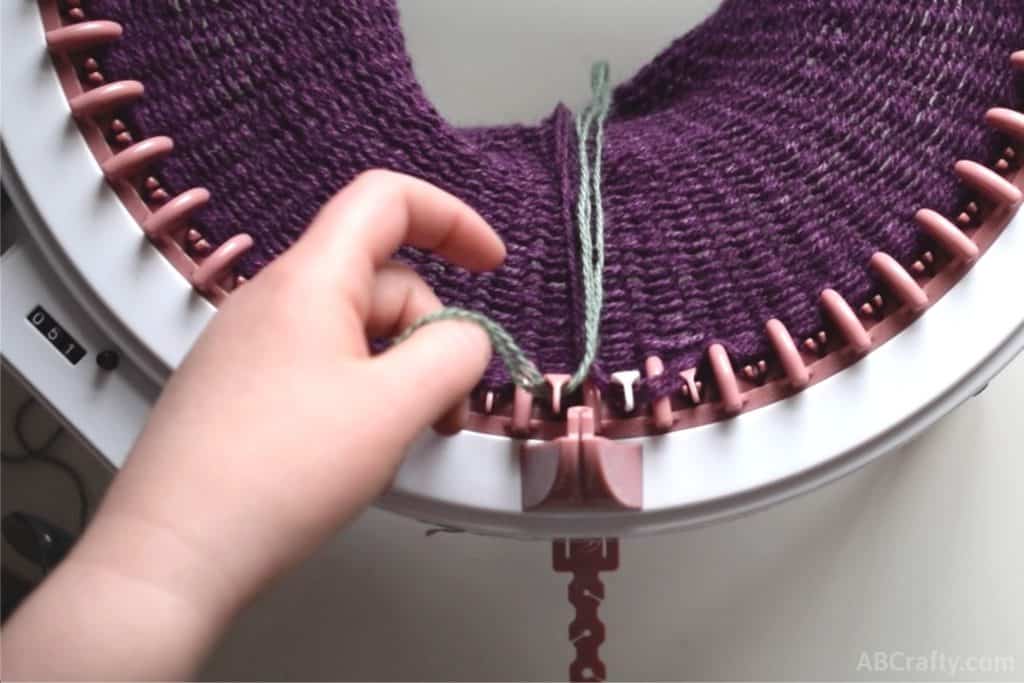 purple yarn hanging in the middle of the knitting with the white needle of the sentro knitting machine to the right and the end of new green yarn starting in front of the next peg to the left