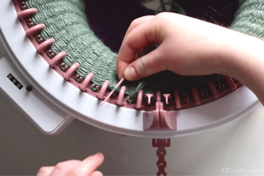 Pink plastic needle going through a loop of green yarn to the left of the yarn holder on the sentro knitting machine