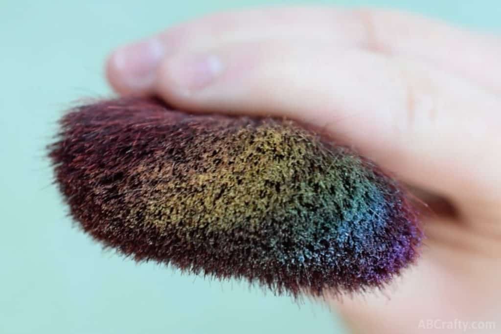 holding a big fluffy makeup brush with rainbow makeup across the brush