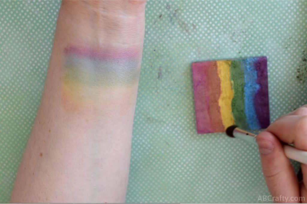 wrist with rainbow makeup on it while dipping a makeup brush into a diy rainbow highlighter