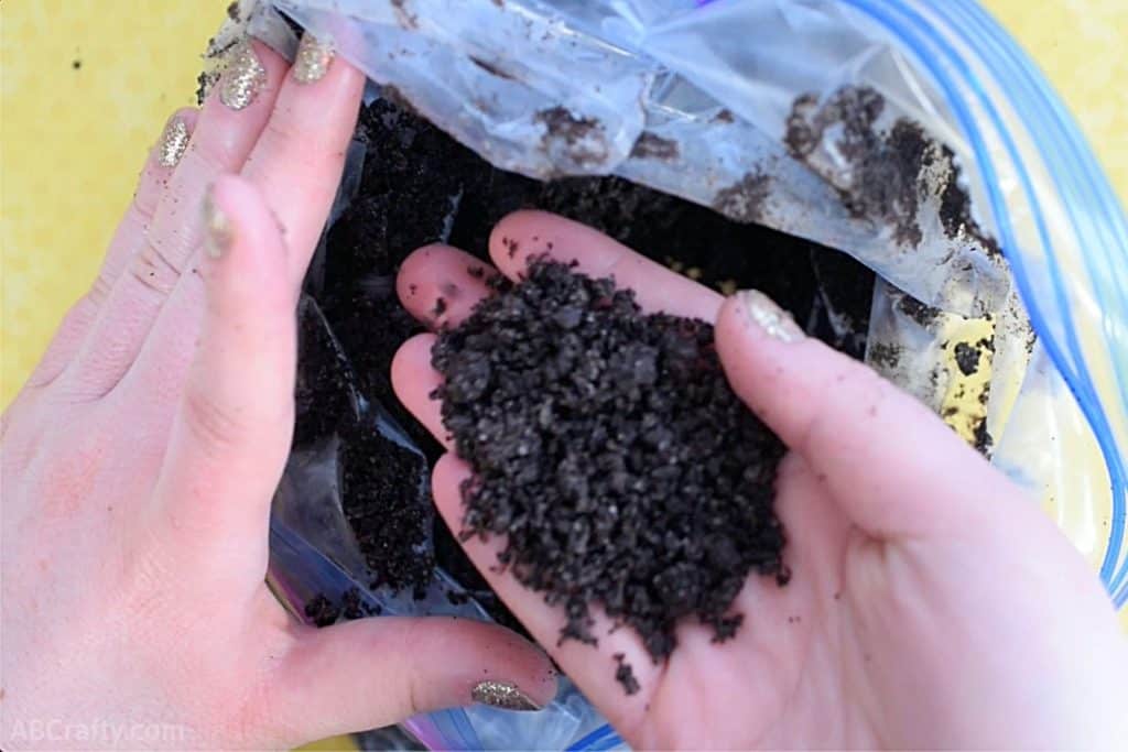 holding a handful of crushed oreos that look like dirt above of a plastic bag filled with the rest of the oreo dirt