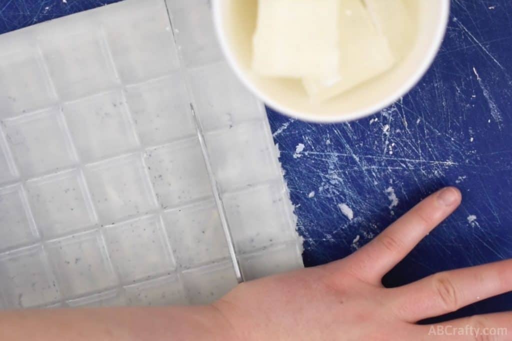 using a knife to cut into clear glycerin soap base with a paper cup next to it that's filled with chunks of clear glycerin soap