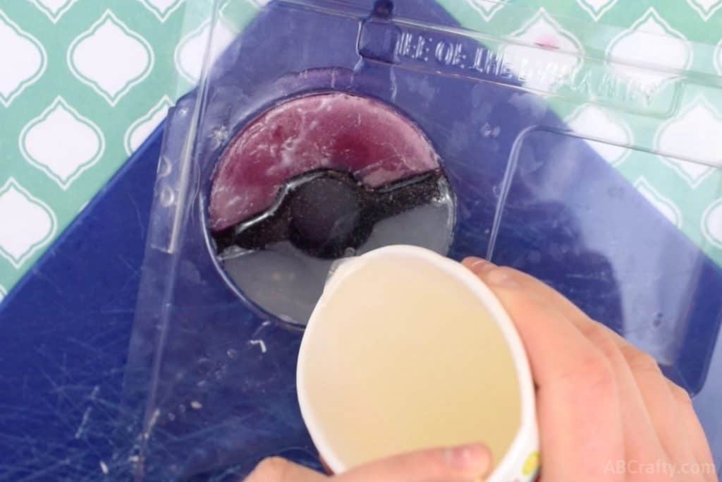 pouring clear soap base over a pokeball shaped soap in a circular soap mold
