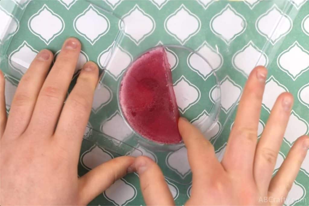 wiping rubbing alcohol on the inside of a half circle of red soap inside a clear plastic round soap mold