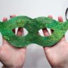 holding a finished green masquerade mask made out of wet felted handmade felt with black satin ribbon hanging from the sides