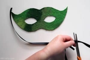 cutting black satin ribbon in front of a green felt mask