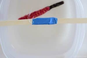 pink and orange pipe cleaner popsicle hanging from two popsicle sticks taped together and placed on top of a plastic container filled with clear liquid