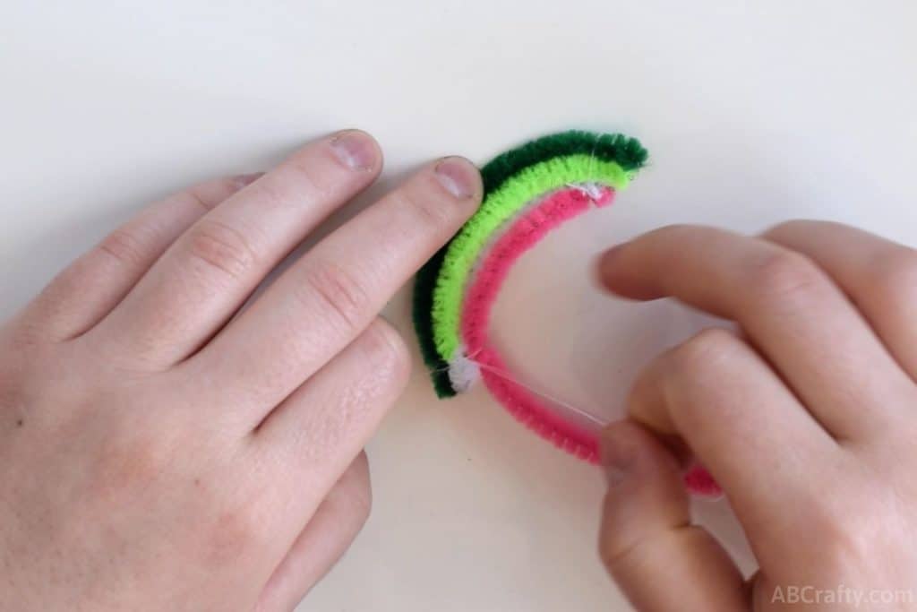 using white thread to tie together the ends of a dark green, light green, white, and pink pipe cleaners that are slightly curved
