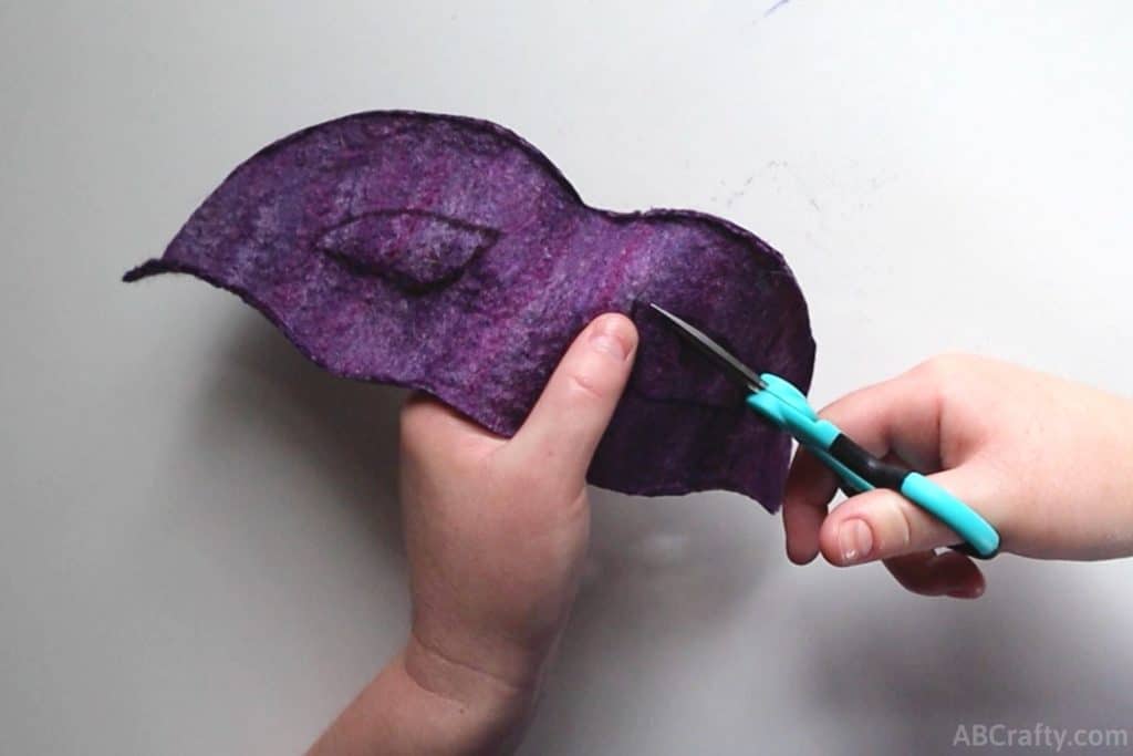 using tiny scissors to cut the outline of an eye hole for a masquerade mask out of handmade purple felt fabric