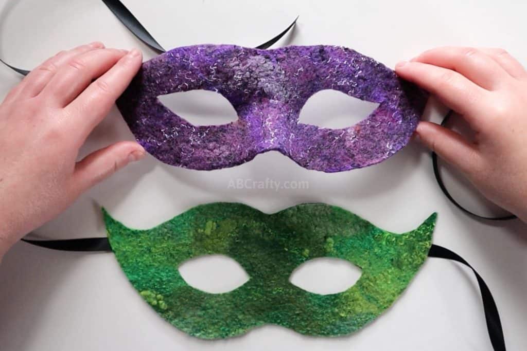 holding the finished handmade purple wet felted Venetian mask above a flat green felted masquerademask