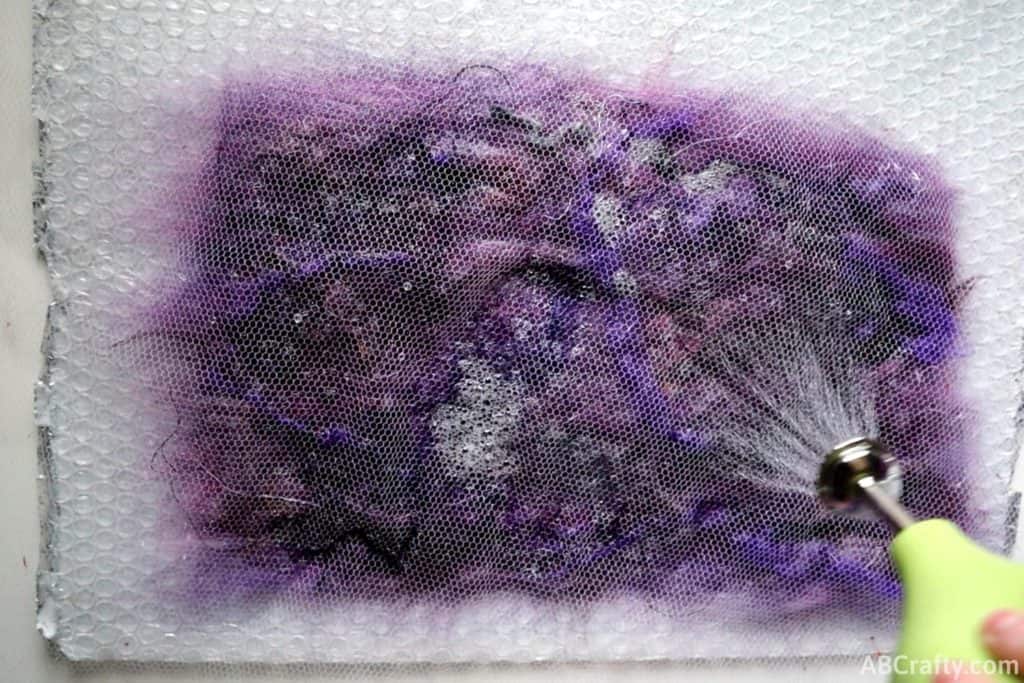 using a ball brasse to spray water onto purple wool and other felting embellishments covered in a netting