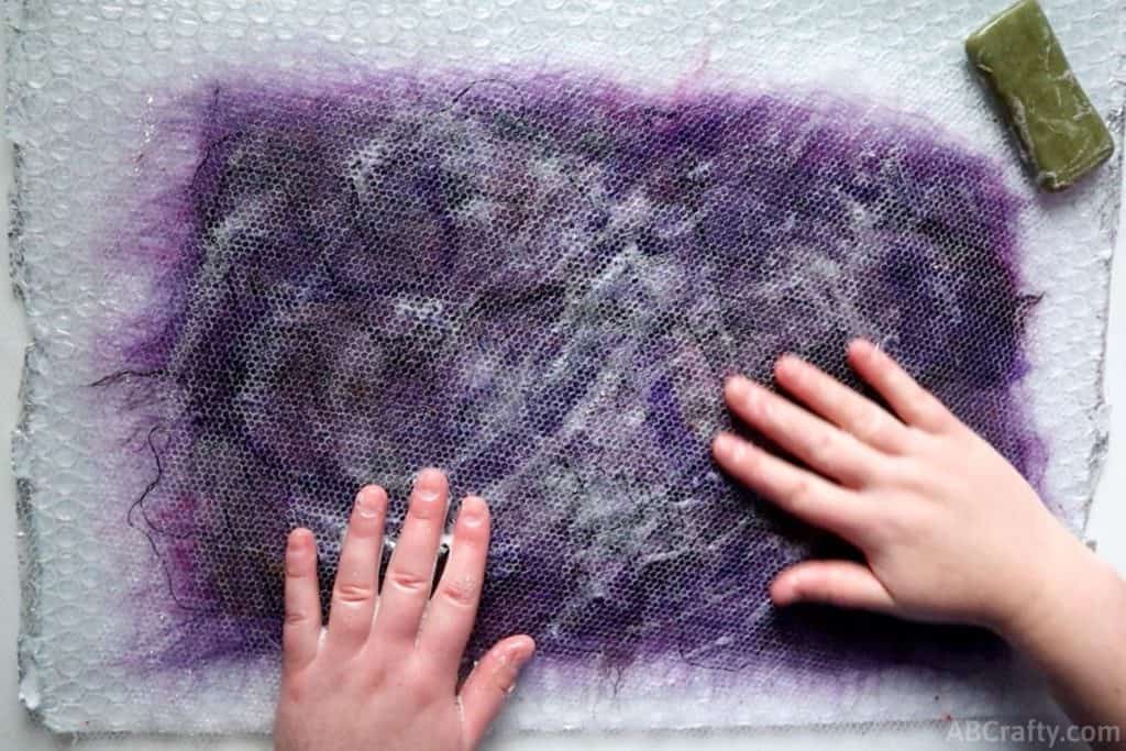 using hands to rub soap and water on top of netting with purple wool and fibers underneath with olive oil soap on the side.